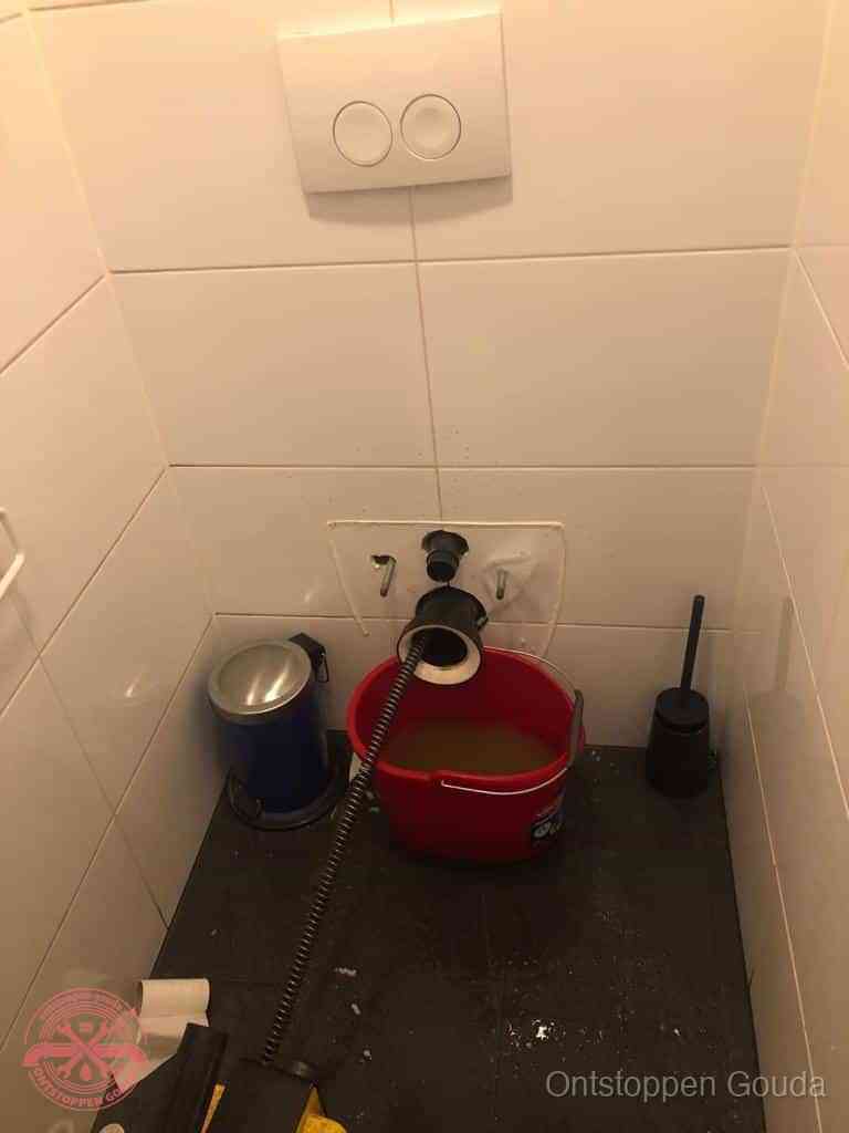 Een ontstopte wand-wc met verwijderde toiletpot waar een ontstoppingsveer in de wanduitgang verdwijnt, het achterhalen waar de verstopping zit in Gouda