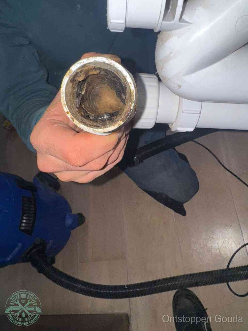 Een hand houdt een losgekoppeld PVC-afvoerbuisstuk vast dat van binnen volledig is dichtgeslibd met vet, vuil en aanslag, de meest voorkomende oorzaak van een verstopte afvoer in Gouda