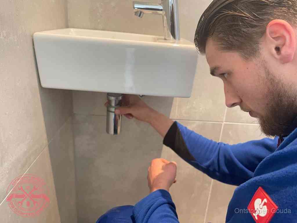 Een monteur draait met de hand de zwanenhalssifon onder een fonteinwasbak in de toiletruimte los om de bron van rioollucht in Gouda te vinden