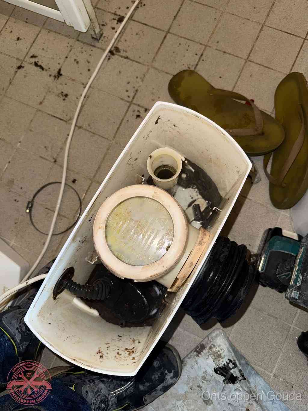 Een gedemonteerde toiletpot op de vloer met ontstoppingsveer en gereedschap ernaast, het verwijderen van een vastzittend voorwerp uit de wc in Gouda