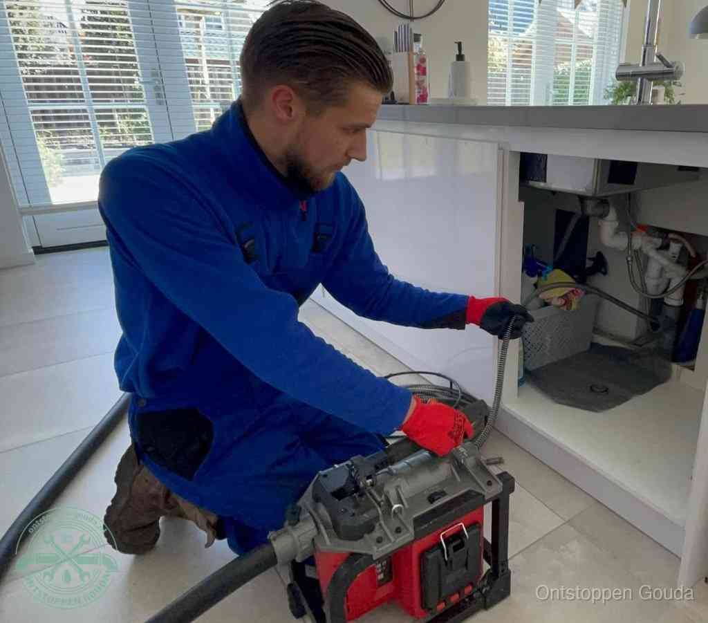 Een monteur met rode handschoenen knielt in een keuken en voert een elektrische ontstoppingsveer in tijdens het professioneel afvoer ontstoppen in Gouda