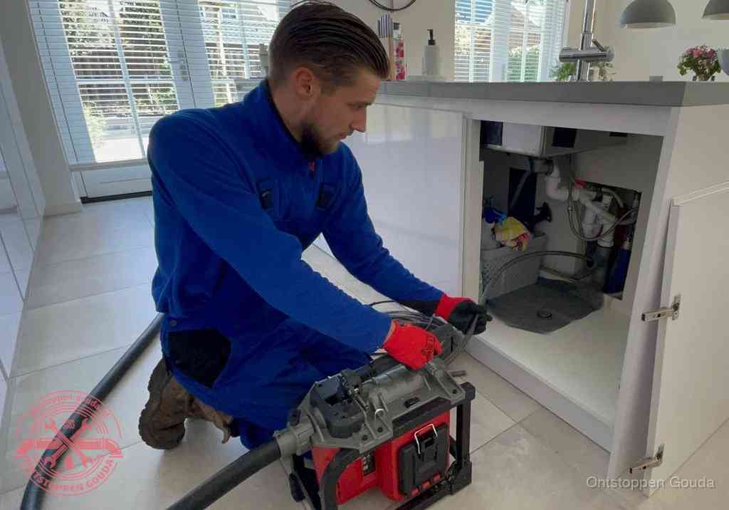 Een monteur voert een professionele elektrische ontstoppingsmachine in de afvoer onder de spoelbak tijdens het gootsteen ontstoppen in Gouda